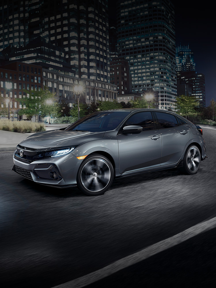 Front driver-side view of the 2021 Honda Civic Sport Touring Hatchback, in Sonic Gray Pearl, driving in city at night.